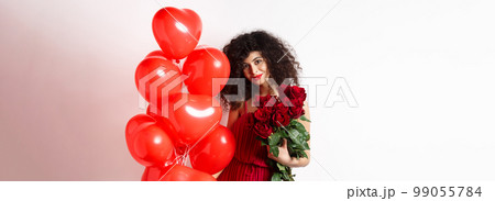 Gorgeous curly haired girlfriend in evening dress, having a date, holding red roses from boyfriend and posing near romantic hearts balloons, white background 99055784