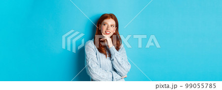 Thoughtful teenage girl with red hair, looking upper right corner logo and thinking, imaging something, standing over blue background 99055785