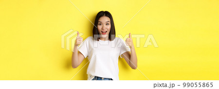 Pretty young asian woman in white t-shirt, showing thumbs up and smiling, praise good offer, recommend product, standing satisfied over yellow background 99055865