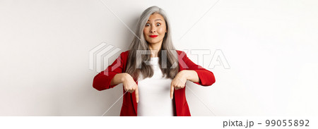 Beautiful asian senior woman in elegant red blazer, pointing fingers down and smiling excited, showing advertisement, white background 99055892
