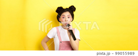 Cute asian teen girl with bright makeup, singing in microphone karaoke, standing against yellow background 99055989