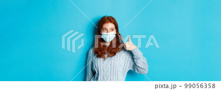 Winter, covid-19 and social distancing concept. Satisfied young redhead woman in face mask showing alright, thumb up gesture and looking left at promo, blue background Winter, covid-19 and social distancing concept. Satisfied young redhead woman in face mask showing alright, thumb up gesture and looking left at promo, blue background 99056358