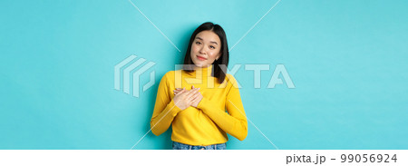 Portrait of beautiful heartfelt woman holding hands on heart, smiling and listening compassionate, standing over blue background in yellow pullover Portrait of beautiful heartfelt woman holding hands on heart, smiling and listening compassionate, standing over blue background in yellow pullover 99056924