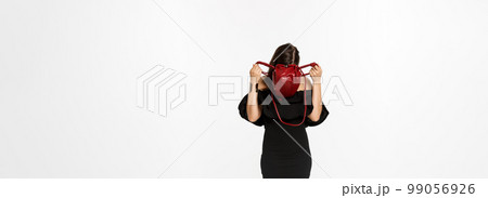Beauty and fashion concept. Full length of young woman sticking head inside purse and searching something, wearing black dress and high heels, standing over white background 99056926