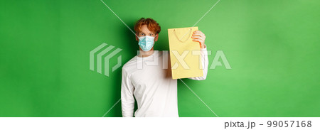Covid-19 and pandemic concept. Surprised man in face mask showing shopping bag and looking at camera, standing over green background Covid-19 and pandemic concept. Surprised man in face mask showing shopping bag and looking at camera, standing over green background 99057168