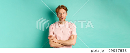 Intrigued redhead guy listening gossips, looking amazed and interested at camera, standing over turquoise background in casual t-shirt 99057638
