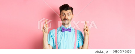 Hopeful young man making wish, holding fingers crossed and smiling, waiting for results with optimism, standing over pink background 99057639