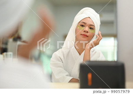 Charming millennial woman in bathrobe applying patches under eyes. Beauty treatment, skincare and cosmetics concept Charming millennial woman in bathrobe applying patches under eyes. Beauty treatment, skincare and cosmetics concept 99057979