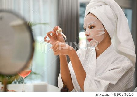 Image of woman with sheet face mask applying drop of essential oil on back of hand, doing her everyday routine at home 99058010