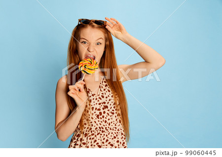Portrait of emotional young beautiful redhaired girl with long glossy hair tasting lollipop and looking at camera isolated over light blue background. 99060445