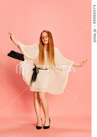 One young beautiful redhaired girl with long hair dancing, posing isolated over light pink background. Concept of positive emotions, happy mood, fashion, youth 99060474