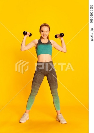 Strength. Happy beautiful girl in sports fitness clothes doing exercises with equipment over yellow background. Sport, fitness, happy positive emotions and active lifestyle concept. Strength. Happy beautiful girl in sports fitness clothes doing exercises with equipment over yellow background. Sport, fitness, happy positive emotions and active lifestyle concept. 99060509