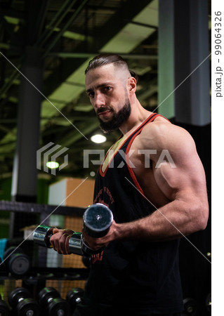A handsome muscular man with a beard in a black T-shirt holds dumbbells in his hands. Sports, healthy lifestyle A handsome muscular man with a beard in a black T-shirt holds dumbbells in his hands. Sports, healthy lifestyle 99064326