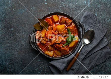 Beef meat stew with potatoes, carrot and delicious gravy in black casserole pot with bay leaves and fresh green herbs with spoon on black dark rustic concrete background from above . 99064937