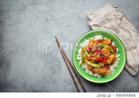 Asian sweet and sour sticky chicken with vegetables stir-fry and rice in ceramic bowl with chopsticks top view on gray rustic stone background, traditional Asian dish. Copy space 99064968