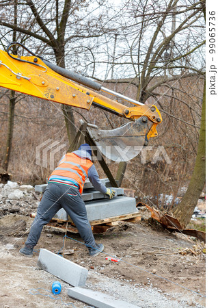 A worker using an excavator bucket unloads pallets of concrete curbs. A worker using an excavator bucket unloads pallets of concrete curbs. 99065736