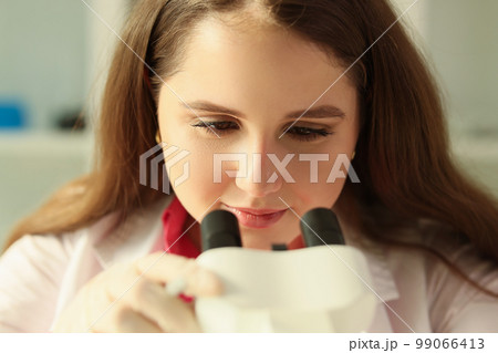 Young female scientist looks through microscope during experiment in research laboratory. Young female scientist looks through microscope during experiment in research laboratory. 99066413