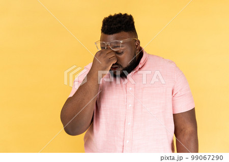 Portrait of frustrated man wearing pink t shirt and optical glasses touching closed eyes, grief or pain after long hours working, feeling hopeless. Indoor studio shot isolated on yellow background. 99067290