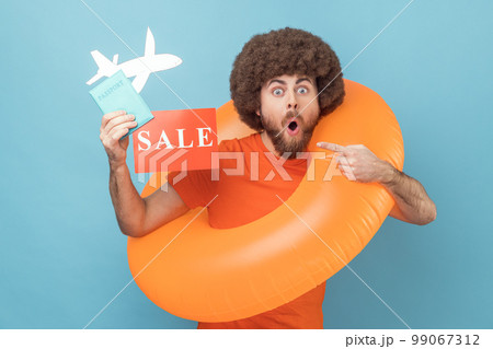 Shocked man with Afro hairstyle standing with orange rubber ring, holding passport document and pointing sale card, tours for low cost. Indoor studio shot isolated on blue background 99067312