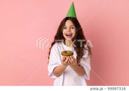 Excited little girl in green party cone on head holding cake with candle and looking at camera with excitement, expressing happiness. Indoor studio shot isolated on pink background. Excited little girl in green party cone on head holding cake with candle and looking at camera with excitement, expressing happiness. Indoor studio shot isolated on pink background. 99067316