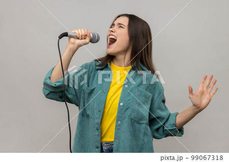 Portrait of woman singing songs in microphone, singer making performance, keeps eyes closed, wearing casual style jacket. Indoor studio shot isolated on gray background. 99067318