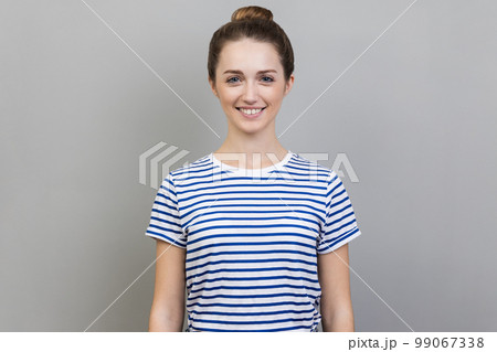 Portrait of attractive charming woman wearing striped T-shirt standing looking at camera with toothy smile, expressing happiness and positive emotions. Indoor studio shot isolated on gray background. 99067338