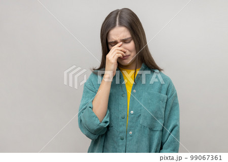 Depressed young woman holding her head down, touching face and crying with dramatic grimace, feeling sorrow regret, wearing casual style jacket. Indoor studio shot isolated on gray background. Depressed young woman holding her head down, touching face and crying with dramatic grimace, feeling sorrow regret, wearing casual style jacket. Indoor studio shot isolated on gray background. 99067361