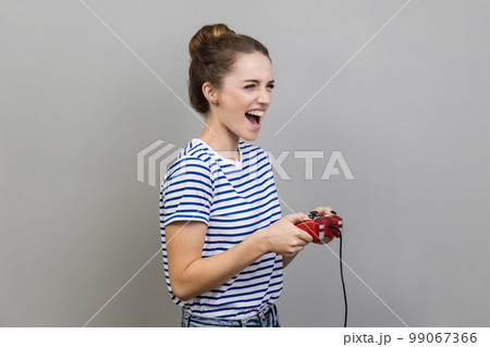 Portrait of woman wearing striped T-shirt holding gamepad, winning video games competition, looking away with happy expression. Indoor studio shot isolated on gray background. Portrait of woman wearing striped T-shirt holding gamepad, winning video games competition, looking away with happy expression. Indoor studio shot isolated on gray background. 99067366