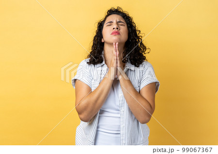 Portrait of woman with dark wavy hair standing praying up heartily, feeling guilty, pleading begging help from heaven, asking apology. Indoor studio shot isolated on yellow background. Portrait of woman with dark wavy hair standing praying up heartily, feeling guilty, pleading begging help from heaven, asking apology. Indoor studio shot isolated on yellow background. 99067367