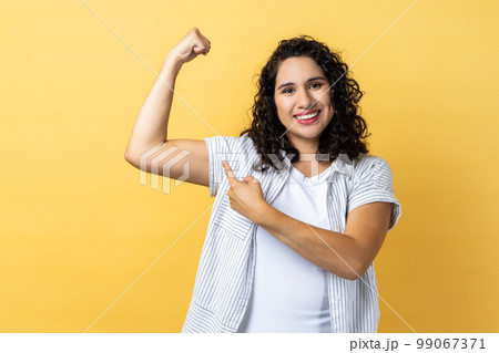 Portrait of woman with dark wavy hair showing her arm muscle and point it with finger, proud of her strength and leadership skills. Indoor studio shot isolated on yellow background. 99067371