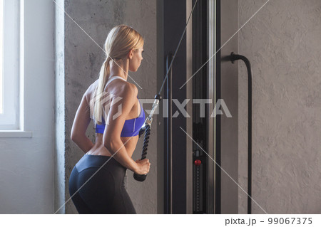 Side view portrait of athletic woman exercising muscles on exercise machine in modern gym, training her triceps, having workout for her arms, wearing blue sporty top and black leggins. Indoor shot. 99067375