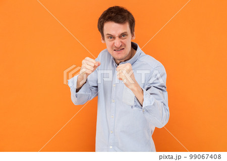 Portrait of angry aggressive man standing clenched fists and being ready to attack, fighting, looking at camera, wearing light blue shirt. Indoor studio shot isolated on orange background. 99067408