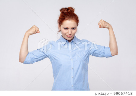 Portrait of strong confident young adult ginger woman wearing blue shirt raising arms showing biceps, female strength and feminism. Indoor studio shot isolated on gray background. Portrait of strong confident young adult ginger woman wearing blue shirt raising arms showing biceps, female strength and feminism. Indoor studio shot isolated on gray background. 99067418
