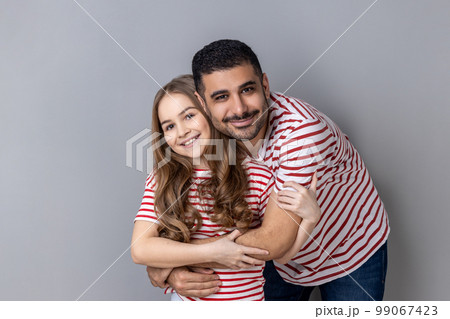 Portrait of loving happy father and daughter in striped T-shirts standing together, dad hugging cute little girl, looking at camera with smile. Indoor studio shot isolated on gray background. Portrait of loving happy father and daughter in striped T-shirts standing together, dad hugging cute little girl, looking at camera with smile. Indoor studio shot isolated on gray background. 99067423