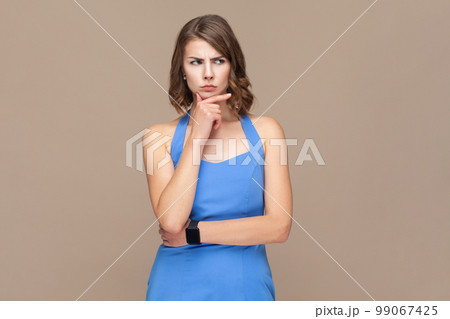 Portrait of attractive thoughtful puzzled woman with wavy hair holding chin, looking away with pensive expression, planning, wearing blue dress. Indoor studio shot isolated on light brown background. Portrait of attractive thoughtful puzzled woman with wavy hair holding chin, looking away with pensive expression, planning, wearing blue dress. Indoor studio shot isolated on light brown background. 99067425