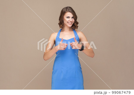 Portrait of attractive positive optimistic woman with wavy hair standing with raised thumbs up, recommended service, wearing blue dress. Indoor studio shot isolated on light brown background. 99067479