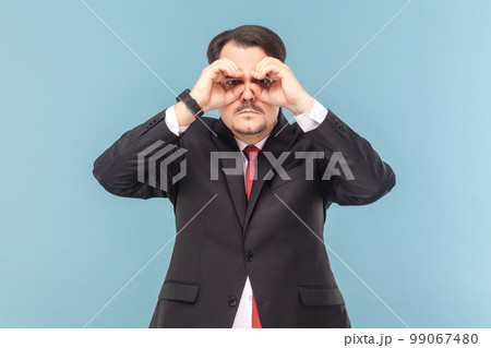 Portrait of serious concentrated man with mustache looking at camera through binocular making with hands, wearing black suit with red tie. Indoor studio shot isolated on light blue background. Portrait of serious concentrated man with mustache looking at camera through binocular making with hands, wearing black suit with red tie. Indoor studio shot isolated on light blue background. 99067480