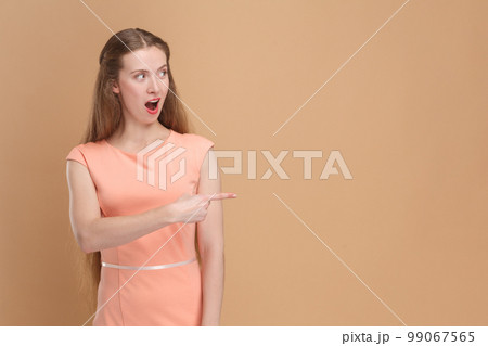 Portrait of shocked amazed surprised woman with long hair pointing aside at advertisement area, copy space for promotion, wearing elegant dress. Indoor studio shot isolated on brown background. 99067565