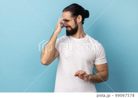 Bad smell. Portrait of man wearing white T-shirt standing pinching her nose with fingers to hold breath, disgusted by stinky intolerable smell. Indoor studio shot isolated on blue background. 99067578