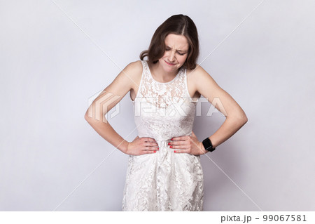 Portrait of sick unhealthy dark haired woman wearing white dress having terrible stomachache, touching her belly, frowning her face. Indoor studio shot isolated on gray background. 99067581