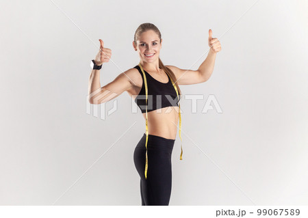 Portrait of powerful muscular athletic blonde woman showing thumbs up at camera and smiling, recommend gym, wearing black fitness clothing. Indoor studio shot isolated on gray background. 99067589