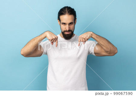 Here and now. Portrait of serious strict man with beard wearing white T-shirt pointing down and looking with arrogance, demonstrating authority. Indoor studio shot isolated on blue background. 99067590