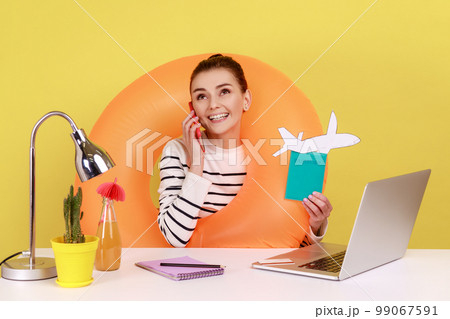 Positive woman with rubber ring, holding card with sale inscription, passport and paper plane, talking phone with happy look. Indoor studio studio shot isolated on yellow background. Positive woman with rubber ring, holding card with sale inscription, passport and paper plane, talking phone with happy look. Indoor studio studio shot isolated on yellow background. 99067591