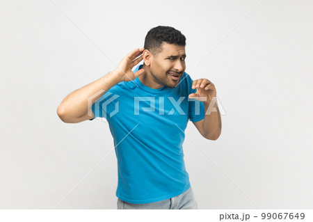 Portrait of unshaven man wearing blue T- shirt standing with frowning face, keeps hand near ear, asking to say louder, trying to hear something silent. Indoor studio shot isolated on gray background. Portrait of unshaven man wearing blue T- shirt standing with frowning face, keeps hand near ear, asking to say louder, trying to hear something silent. Indoor studio shot isolated on gray background. 99067649