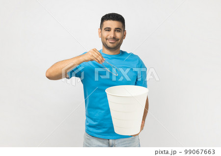 Portrait of satisfied delighted unshaven man wearing blue T- shirt standing after laser eyesight correction, throwing away his eyeglasses. Indoor studio shot isolated on gray background. 99067663