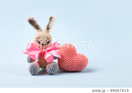 Knitted gray bunny with a crocheted pink heart on a blue background. Happy Valentine's Day, Mother's Day and birthday greeting card. 99067819