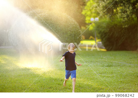 Funny little boy playing with garden sprinkler in sunny city park. Elementary school child laughing, jumping and having fun with spray of water. Summer outdoors activity for kids. 99068242