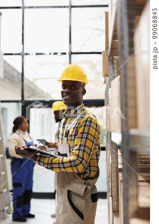 Retail storehouse smiling operator holding clipboard with instructions and looking at camera. African american post office storehouse manager in protective helmet smiling portrait Retail storehouse smiling operator holding clipboard with instructions and looking at camera. African american post office storehouse manager in protective helmet smiling portrait 99068845