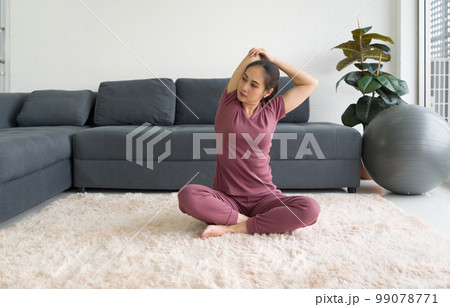 Asian housewife stretching while sitting during home workout on carpet in living room. 99078771