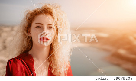 Close up portrait of curly redhead young caucasian woman with freckles looking at camera and smiling. Cute woman portrait in a pink long dress posing on a volcanic rock high above the sea at sunset 99079205